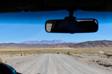 Driving a vehicle along a remote dirt track through the dry Bolivian landscape. In the distance, rugged mountains meet a bright white salt flat under a clear blue sky.