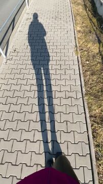 POV walking legs. Shadow of pedestrian walking on sidewalk, elongated silhouette cast across interlocking pavers camera viewpoint from chest. Video taken on mobile device.