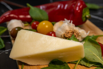 a wedge of hard cheese resting on a wooden cutting board, surrounded by fresh Italian cooking ingredients including cherry tomatoes, garlic cloves, baby spinach, and a large red pepper.