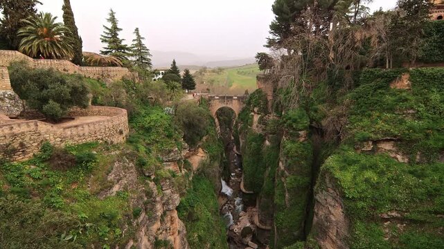 Cinematic wide shot of the historic Puente Viejo bridge crossing the steep Tajo de Ronda canyon with lush greenery and river below in Andalusia, Malaga, Spain.
