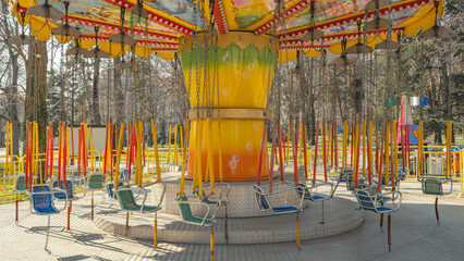 Colorful chain carousel ride at an amusement park. A bright yellow and orange swing ride stands empty in a sunlit park surrounded by trees © John_Doo78