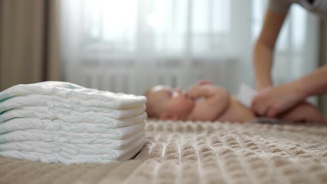 Pile Of Diapers Beside Baby During Diaper Change, White Newborn On Soft Bed, Caregiver Hands Preparing Wipes, Clean Hygiene Routine, Bright Window Light, Calm Nursery Supplies Visible