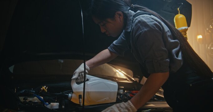 Young Asian Female Mechanic in Work Overalls Carefully Pours Washer Fluid in Car Under Open Hood. In Garage, Girl Focuses on Precise Maintenance, Ensuring Optimal Vehicle Performance and Reliability.