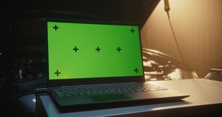 Laptop With Green Screen and Black Tracking Points, Ideal For Motion Tracking and Screen Compositing, Positioned Next to Open Car Hood in Sun Drenched Workshop. Post Production and Digital Marketing.
