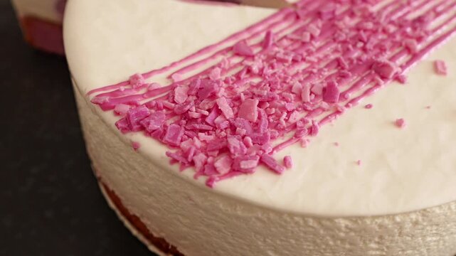 A frosted cake sits on a black plate. It features white icing, diagonal pink drizzle, and bright pink sprinkles.