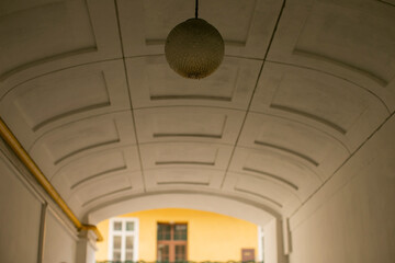 Fototapeta premium Arched passageway with vaulted ceiling, hanging globe light, warm yellow courtyard beyond, soft glow, textured stucco, urban detail