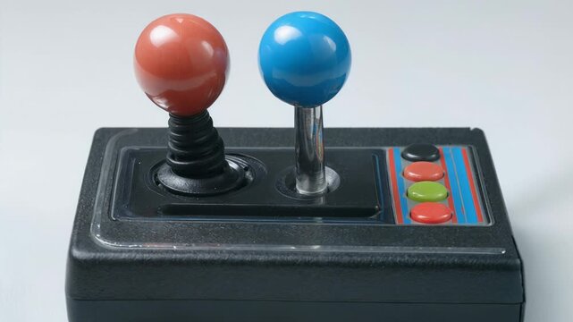 A retro-style black plastic arcade joystick controller with dual ball-top sticks in red and blue and multicolored action buttons sits on a light grey surface in a close-up studio composition.