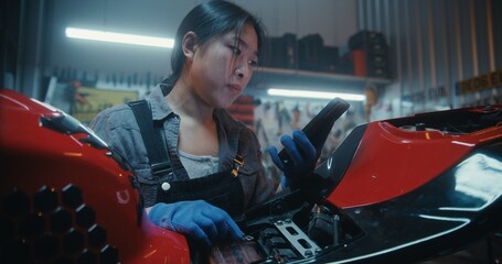 Young Woman Repairing Motorcycle in Modern Garage Uses Smartphone With Voice AI Assistant. Girl Describes Wiring Issues, Listens to Step By Step Instructions, Reconnecting According Digital Guidance. © Framestock