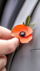 Obraz premium Hand pinning a vivid red poppy boutonniere with green leaves onto a grey suit lapel, close up symbolizing remembrance, tribute and respect for veterans and fallen soldiers