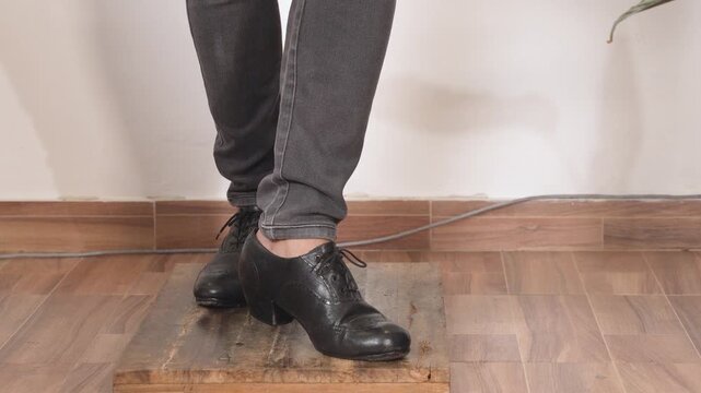 Cropped view of a person wearing black leather dance shoes and gray pants on an indoor floor, making a flamenco dance steps on a wooden platform.