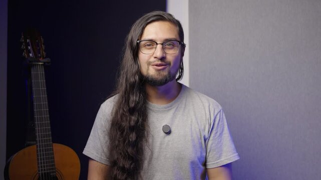 Medium shot of a man with long, wavy dark hair, a goatee, and glasses. He is wearing a simple grey t-shirt with a small lapel microphone attached.