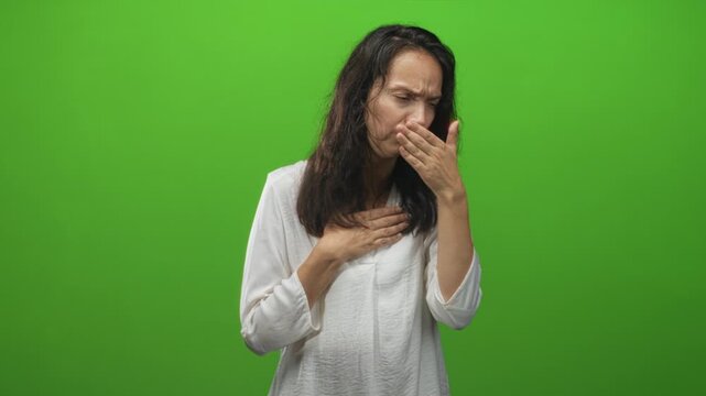 Woman with hand over mouth and hand on chest coughing in studio wearing white blouse against green screen; discomfort.