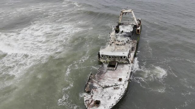 Drone lowers while orbiting around the side of Zeila Shipwreck with waves crashing on overcast day off the coast of Namibia