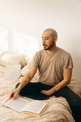 Fototapeta premium Tattooed man with a beard and shaved head sitting cross-legged on a bed, reading an open book in a bright, minimalist room with natural sunlight. Concept of relaxation, peace, and mindful literacy.