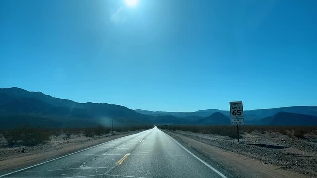 Desert highway with speed limit sign in Death Valley California. Straight asphalt road with a 65 mph speed limit sign leading toward mountains in an arid landscape.