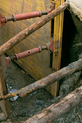 Deep trench with timber braces and yellow shoring, urban excavation site showing damp soil and structural struts, road crew securing support