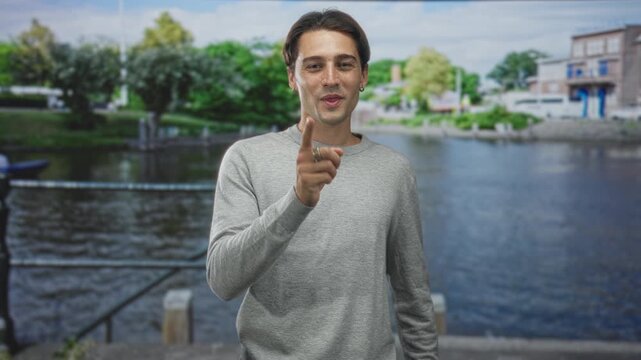 Man pointing finger and gesturing with hands over chest beside a riverside building at a canal quay while addressing the camera; friendly gratitude.