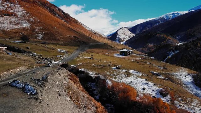 Remote caucasus valley with mountain on which car is driving cabin and winding dirt road, light snow patches near shelter, towering peaks in background, drone perspective serene overlanding basecamp