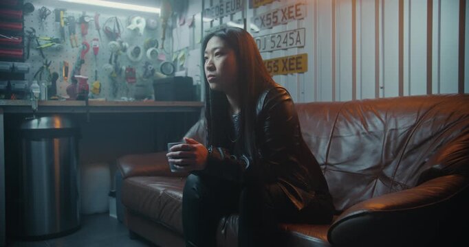 Young Asian Woman in Leather Gear Sits on Couch in Industrial Garage, Holding Mug of Hot Tea Warm Up After High Speed Motorcycle Trip. Motorcycle Lifestyle and Post Ride Recovery Concept. Dolly Shot.
