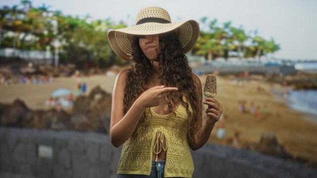 Woman pointing finger at a popsicle while holding it near her chest on a street promenade by a sandy beach with palm trees; curiosity.