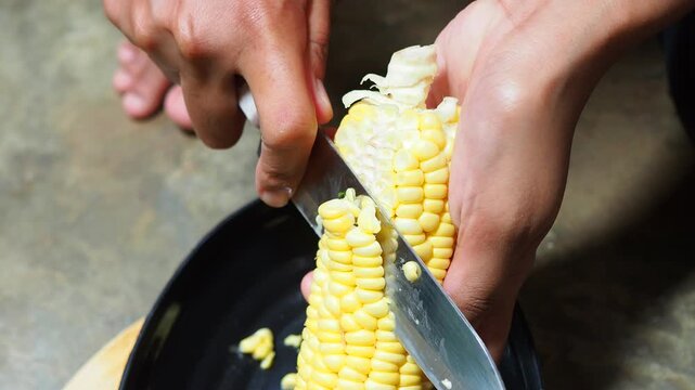 Close-up of hands cutting fresh yellow corn kernels off the cob with a knife into a black bowl