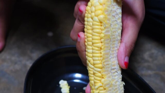 Close-up of hands using a knife to cut and remove fresh yellow corn kernels from a corn cob into a black bowl, preparing ingredients for a meal.