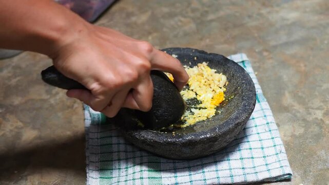 Close-up of a person's hand grinding garlic and spices in a traditional stone mortar and pestle on a checkered cloth, preparing ingredients for cooking.