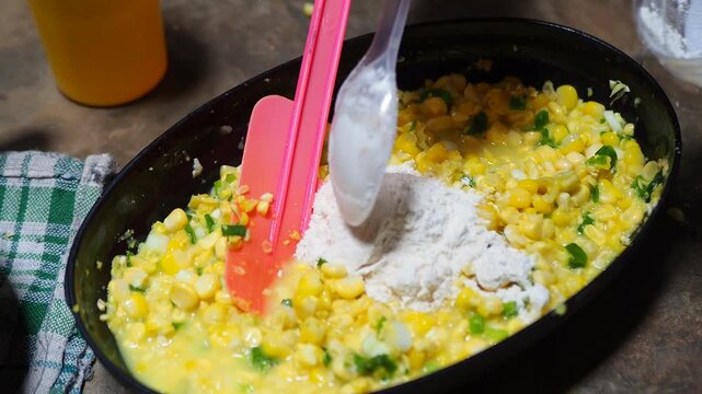 Adding flour to fresh corn kernel mixture with chopped green ingredients in a black bowl for preparing homemade fritters.