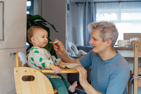 Young father feeding with a spoon adorable messy toddler baby sitting in high chair. Cute baby learning to eat food by himself. Kid enjoying healthy nutrition. family bonding moment during mealtime.