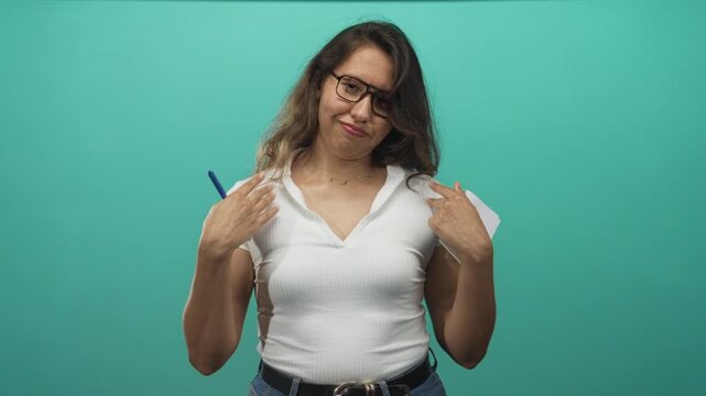 Young hispanic woman in white shirt points fingers to chest while holding pen and notepad in teal studio; sarcasm attitude.