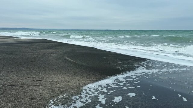 The view shows waves rolling onto a sandy beach under gray skies. The shoreline is calm with no people in sight. Water meets the shore creating froth as waves break.