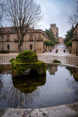 Fototapeta premium Scenic view of a mossy stone fountain reflecting a medieval cathedral tower and historic buildings in a quiet cobblestone square of a spanish village under an overcast sky in winter