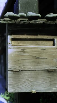 A wooden mailbox stands with stones placed on top in a quiet area. The mailbox is weathered and shows signs of age. The surrounding setting is simple and unadorned, with no people nearby.