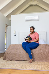 African American woman in pink top sitting on bed in minimalist bedroom reading paperback book