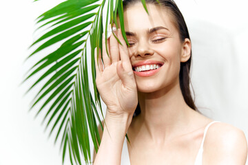 Young woman with a bright smile hiding behind a palm leaf, showcasing natural beauty and tropical...