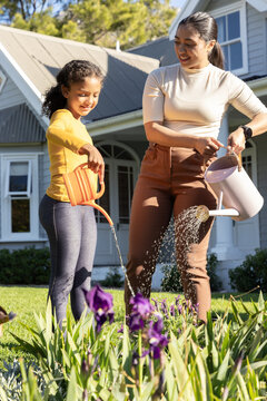 Mother and school-age daughter watering purple iris in yard using orange and white watering cans