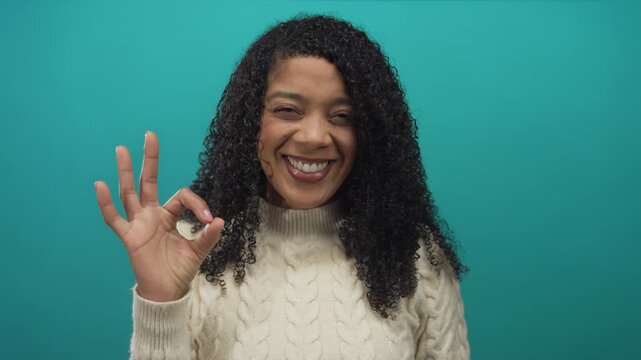 African american woman with curly hair smiles and holds ok sign in studio with teal backdrop; approval.