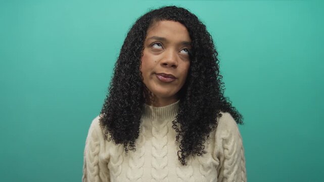 African american woman rolling eyes against teal background in studio wearing cream cable knit sweater; boredom.