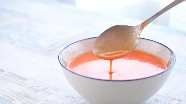 Hand dips a spoon into fresh gazpacho served in a ceramic bowl showing traditional cold soup from Spain prepared with fresh vegetables.