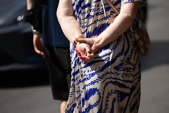 Senior woman's hands clasped behind back, red nails, unposed movement, natural candid motion, hand skin texture, close-up, at Chatuchak Market.