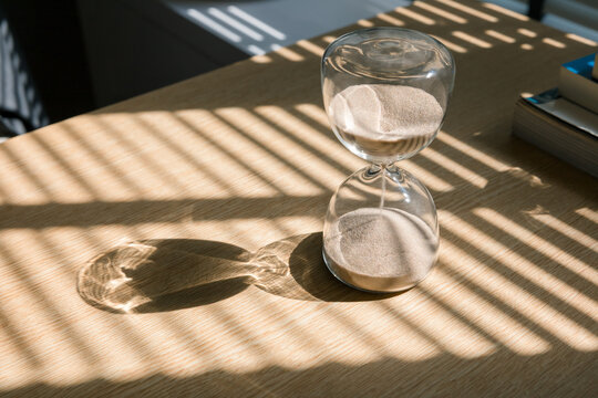 Sand flows through the glass spheres of an hourglass on a wooden desk, casting beautiful shadows in the natural light from the window, measuring the passage of time the concept of timing. 