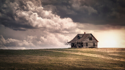 A spooky old, abandoned farmhouse atop a hill in a field of brown grass under a stormy sky.