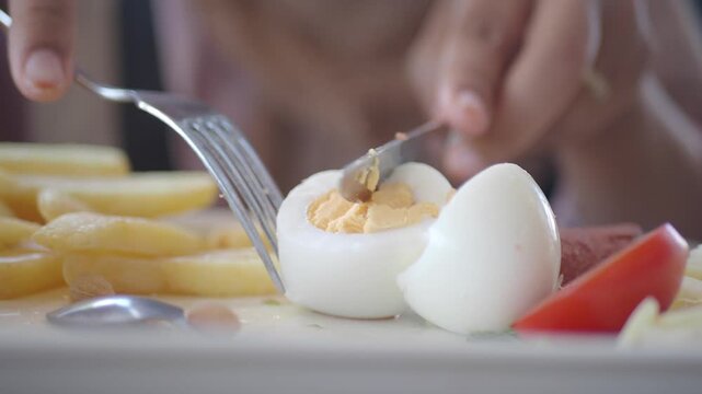Close up of person hands cutting boiled egg into pieces with knife and fork on white plate for breakfast, slow motion cinematic footage