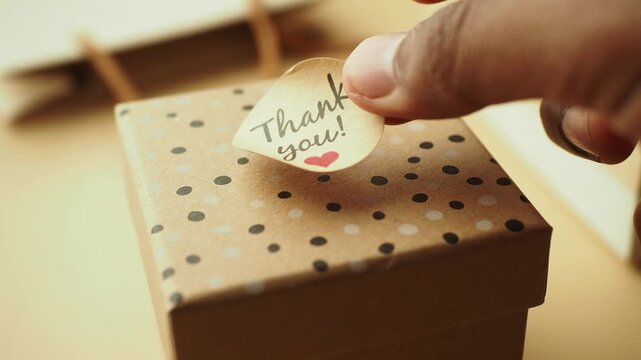 Hands placing thank you sticker on brown polka dot gift box close up view