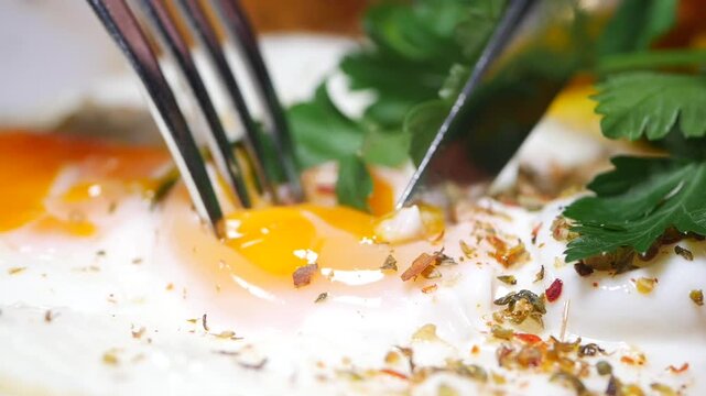 Macro view of knife and fork cutting into runny egg yolk of fried egg with spices and parsley