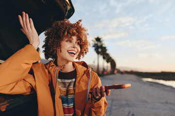 Cheerful woman with curly hair smiling outdoors near car, wearing rainbow sweater and orange...