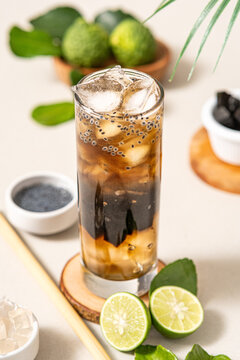 a glass of ice containing nata de coco, grass jelly, ice cubes, basil and grass jelly flavoring, using a long round glass on a round wooden coaster, with a white background.