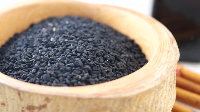 Black cumin seeds in wooden bowl with oil bottle and cinnamon sticks on white background