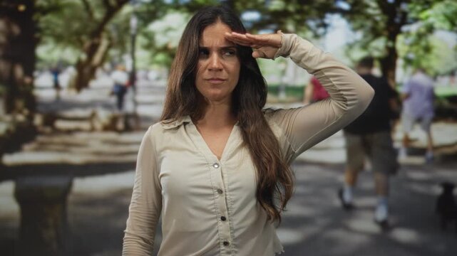 Woman salutes with hand to forehead in city park walkway while facing camera and showing intent face expression; determination.