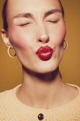Plakat Close-up of a woman with red lipstick puckering lips and closed eyes. She wears hoop earrings and a knitted sweater. Concept of beauty, makeup, and facial expression.